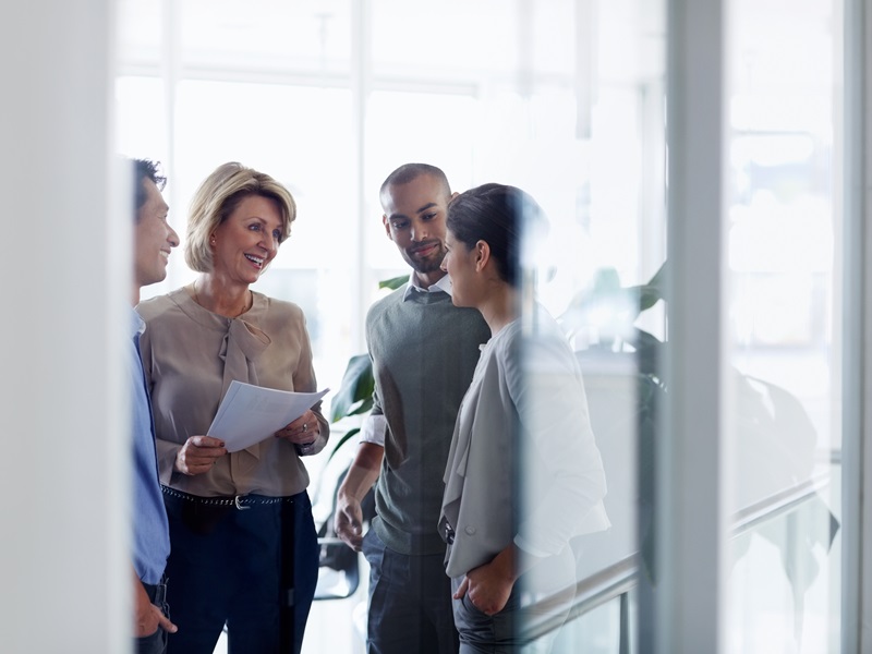 In einem Büro stehen mehrere Personen beieinander. Sie unterhalten sich und lächeln. EIne Dame hält ein paar Blatt Papier in der Hand.